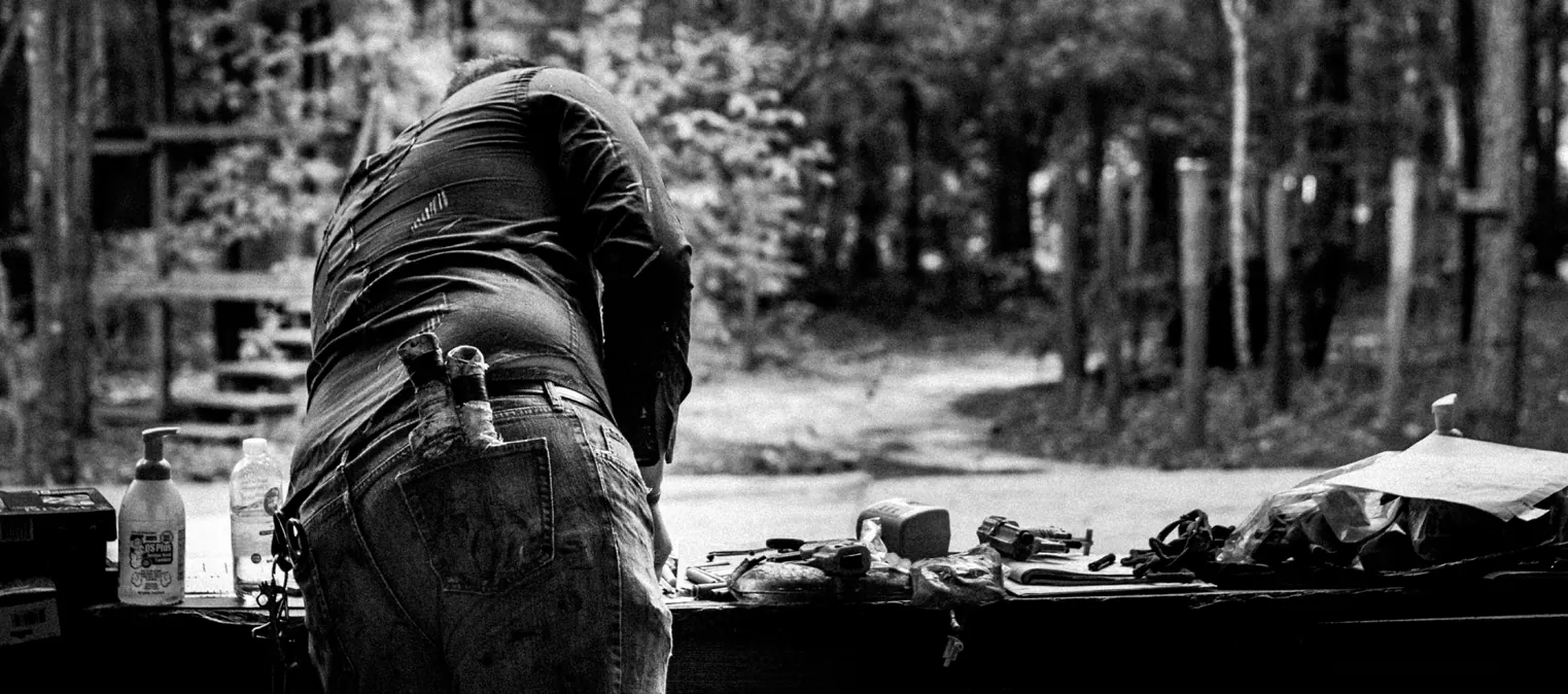 Black-and-white photo of a person in a tactical vest seen from behind, bending over a table outdoors in a wooded camp.