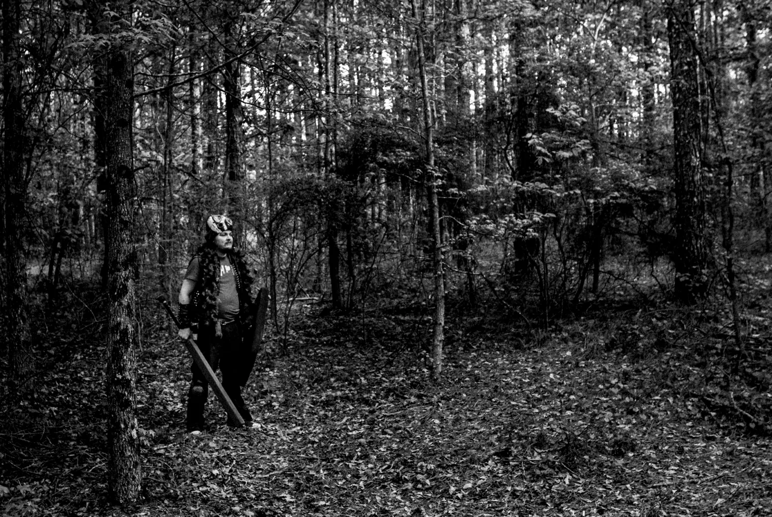 Black-and-white photo of a horned, armored figure with a sword standing among pine trees in a forest.