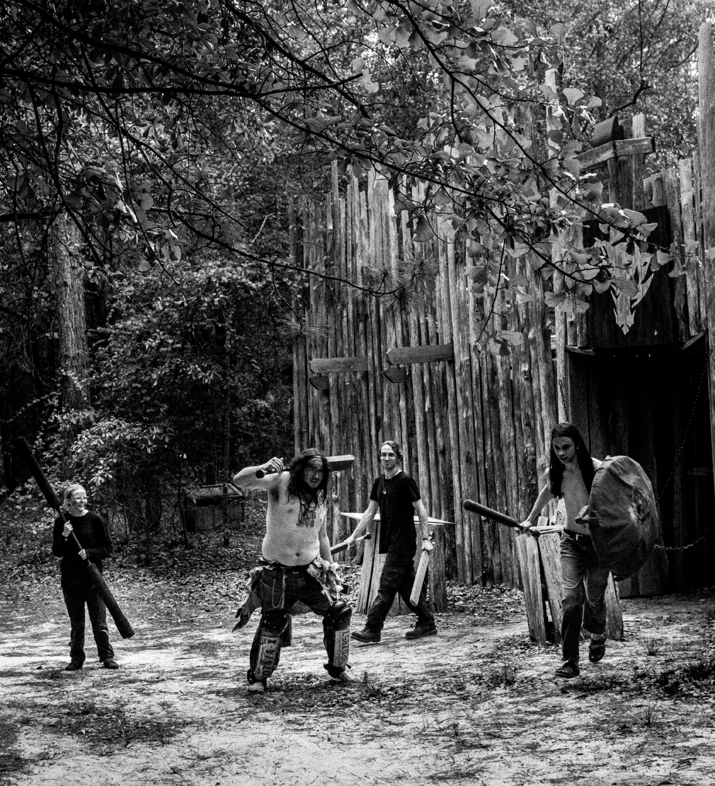 Black-and-white photo of four costumed Khans raiders, armed with clubs and a shield, walking out of a tall log-palisade fortress gate.