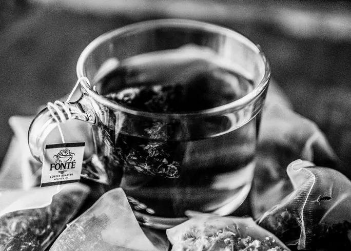 Black-and-white photo of a glass cup of tea with a tea bag label reading “Fonté” hanging over the rim.