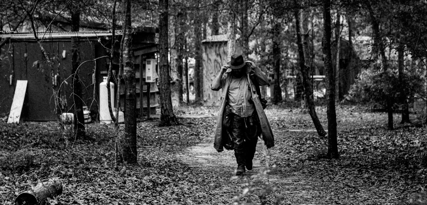Black-and-white photo of a lone wanderer in a long coat and wide-brimmed hat, head bowed, walking down a leaf-covered path in front of a wooden shack.