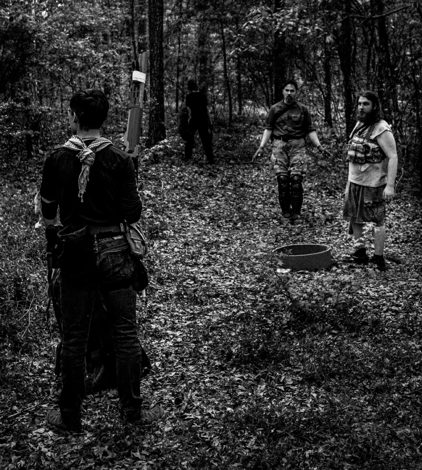 Black-and-white photo of four wastelanders in a leaf-strewn pine forest; one in the foreground, back to the camera, carries a rifle while three others stand facing him in the clearing.