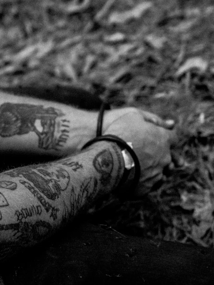 Close-up black-and-white photo of a heavily tattooed forearm and wrist resting on leaf litter.
