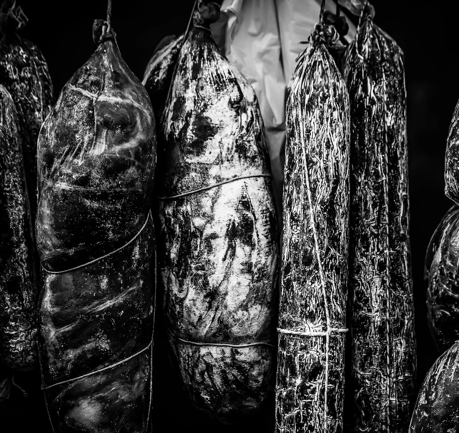 Black-and-white close-up photo of cured sausages hanging in a row, tied with string.