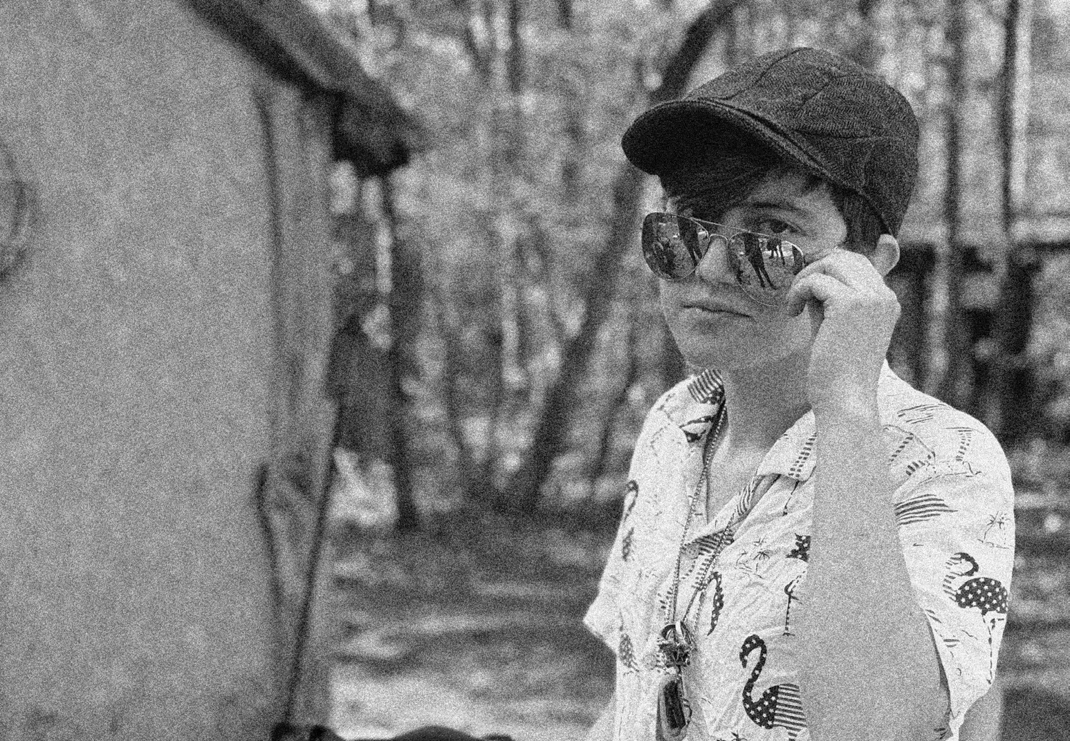 Black-and-white portrait of a person in a flat cap, aviator sunglasses and a flamingo-print shirt, lowering the sunglasses with one hand, in front of a wooden hut.