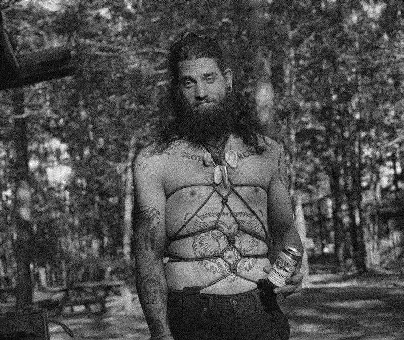 Black-and-white photo of a bearded, tattooed man in a chest harness holding a can, standing in a pine park with picnic tables behind him.