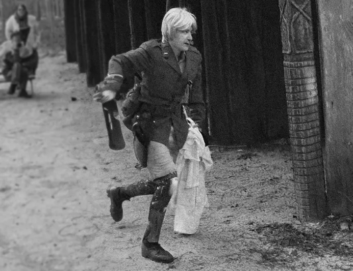 Black-and-white photo of a blonde figure in a jacket and boots running along a dirt track past a wooden palisade, carrying a bottle and a white cloth.