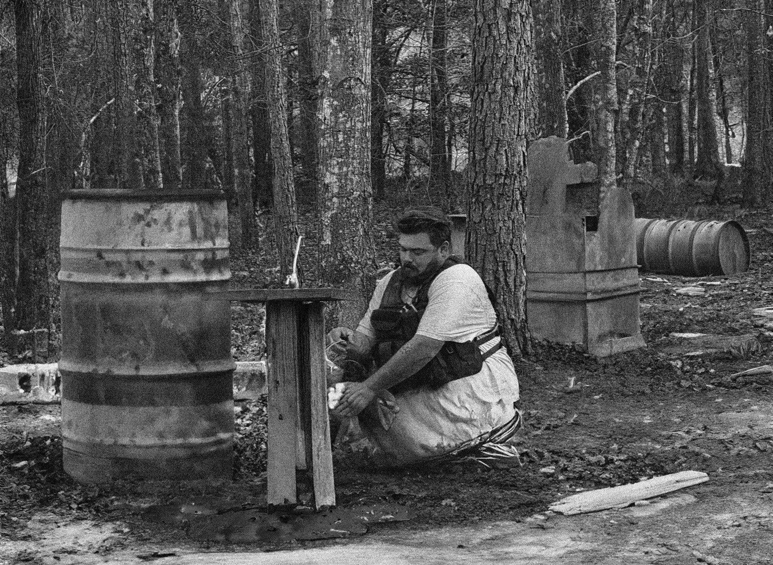 Black-and-white photo of a man in a tactical vest crouching beside a rusty barrel and a wooden stand at a muddy well in a pine forest.